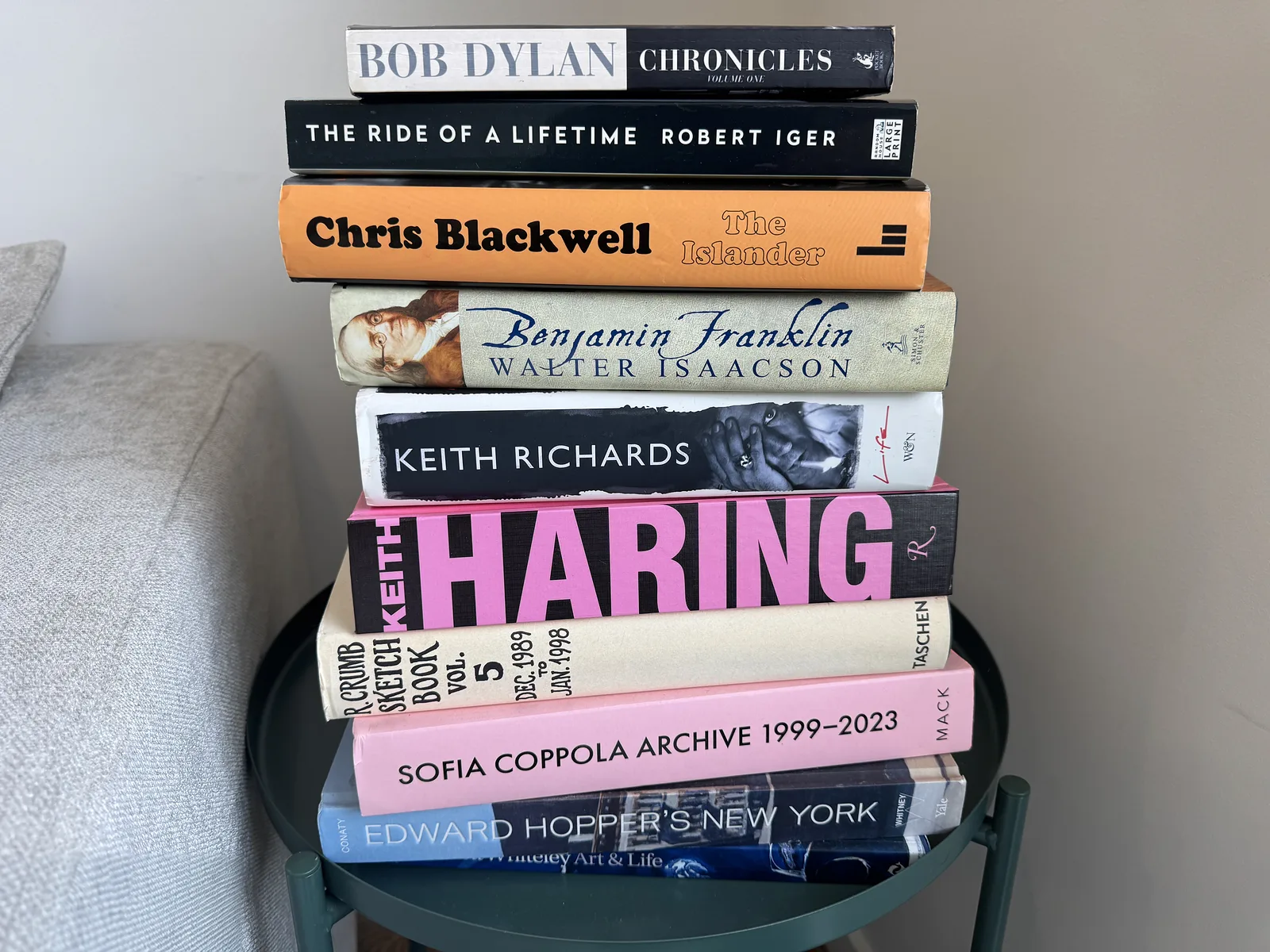 A stack of biographies and art books on a small green side table next to a couch, including Bob Dylan's Chronicles, Robert Iger's The Ride of a Lifetime, Chris Blackwell's The Islander, Walter Isaacson's Benjamin Franklin and Keith Richards' Life
