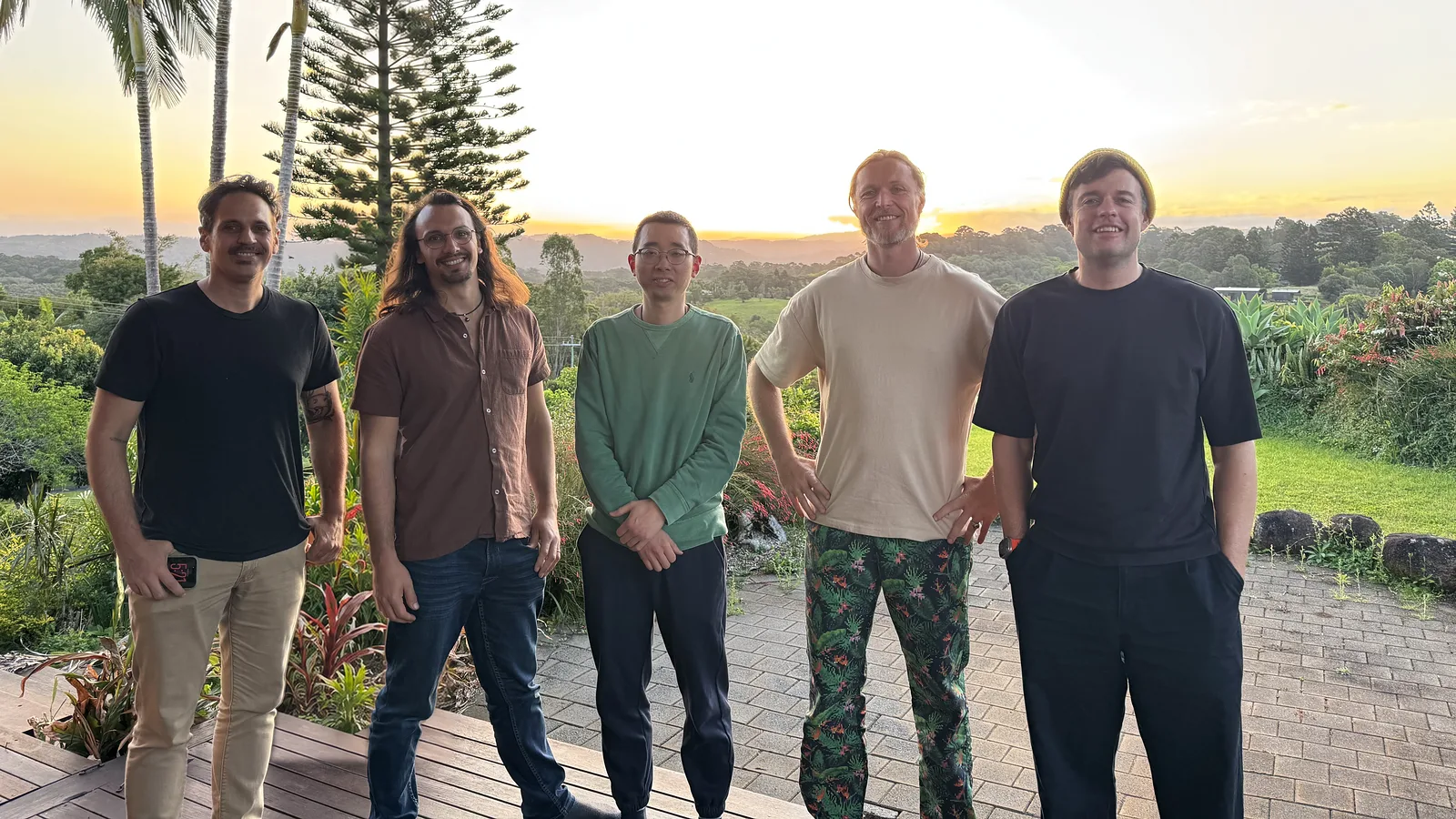 The five-person Nitrosend team standing together on a deck at sunset, with palm trees and the green hills of the Byron Bay hinterland behind them